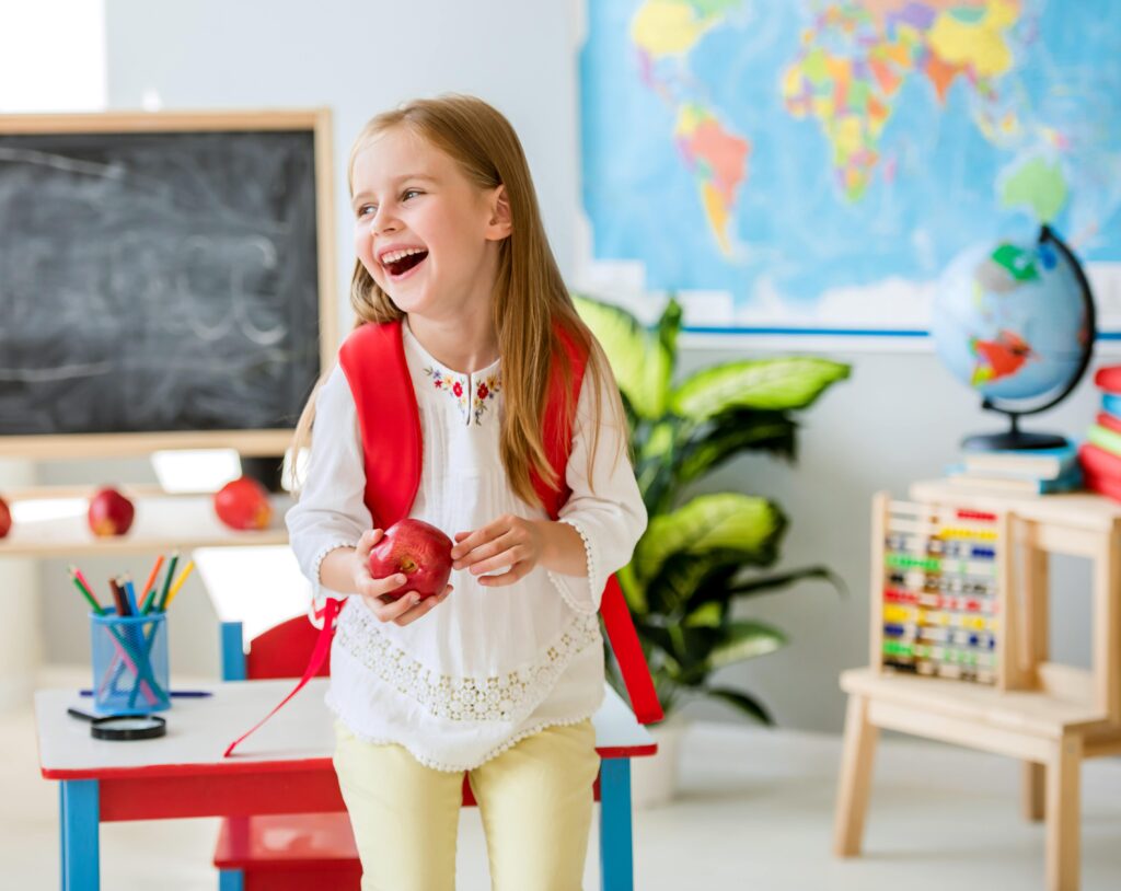 Child with backpack smiling while holding apple in classroom